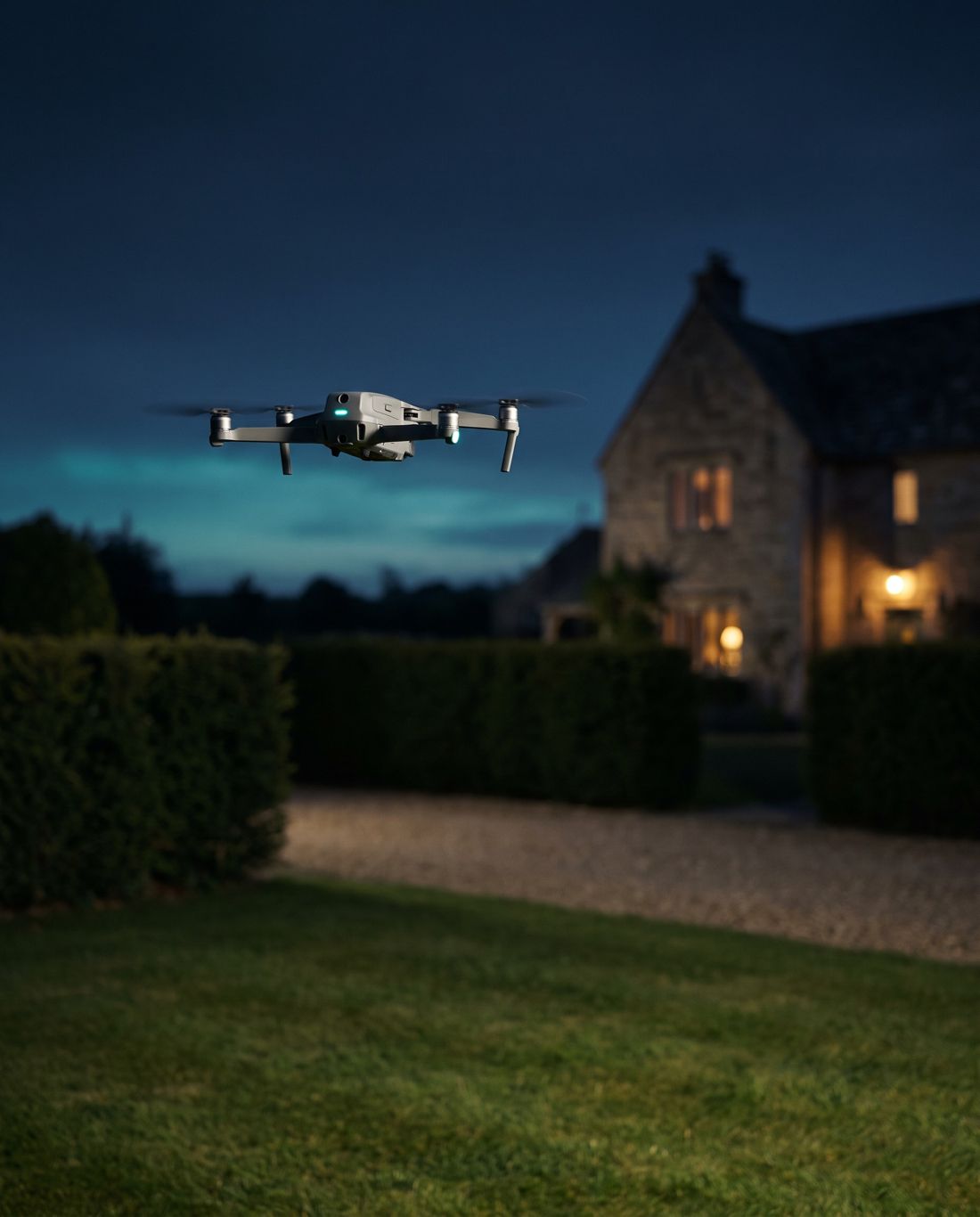 A small grey drone hovering over a lawn at dusk, with clipped hedges and the lit windows of a stone house behind it.